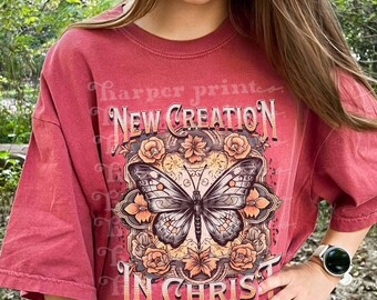 May include: A brick red short-sleeve t-shirt featuring a central graphic of a butterfly surrounded by floral patterns. The text reads NEW CREATION IN CHRIST and 2 Corinthians 5:17. The shirt has a crew neckline and a loose, relaxed fit.