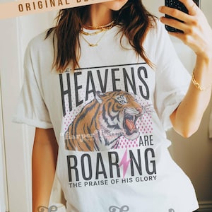 May include: White t-shirt with a tiger graphic and the text "HEAVENS ARE ROARING". The design includes a pink lightning bolt and the phrase "THE PRAISE OF HIS GLORY". The shirt also has the text "harper house THREE SIXTEEN".