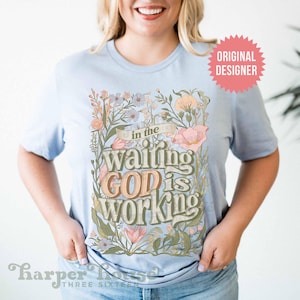 May include: A light blue t-shirt with a floral design and the text "in the waiting GOD is working".