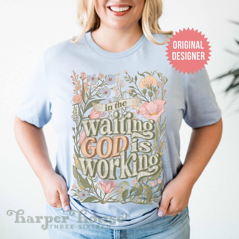 May include: A light blue t-shirt with a floral design and the text "in the waiting GOD is working".