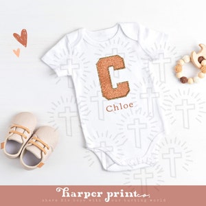 May include: White baby onesie with a large, sparkly, copper-colored letter C and the name Chloe printed below. The onesie is surrounded by a pattern of small crosses.