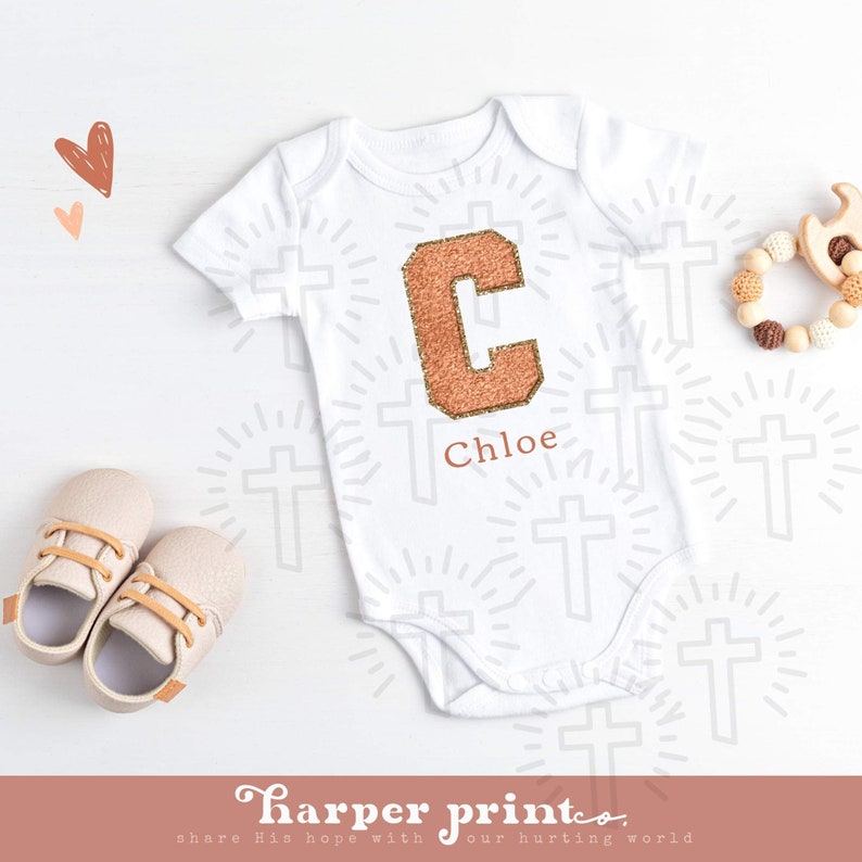 May include: White baby onesie with a large, sparkly, copper-colored letter C and the name Chloe printed below. The onesie is surrounded by a pattern of small crosses.