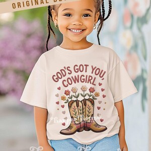 Op de afbeelding: Een cr&egrave;mekleurig T-shirt met de tekst "GOD'S GOT YOU COWGIRL" in een rode, getextureerde letter. De afbeelding bevat cowboylaarzen met bloemen en harten. Het shirt wordt gedragen door een lachend kind.