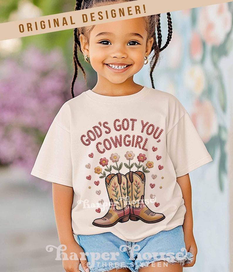 Op de afbeelding: Een cr&egrave;mekleurig T-shirt met de tekst "GOD'S GOT YOU COWGIRL" in een rode, getextureerde letter. De afbeelding bevat cowboylaarzen met bloemen en harten. Het shirt wordt gedragen door een lachend kind.