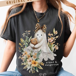May include: Dark gray t-shirt featuring a cartoon ghost holding a Bible, adorned with a leopard print bow and surrounded by floral elements. The text "armed & dangerous" is below the ghost. The shirt also has the text "Harper House Three Sixteen".