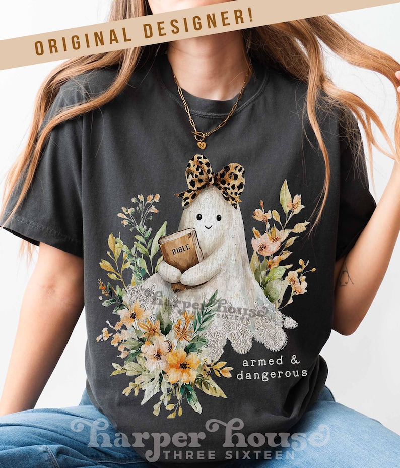 May include: Dark gray t-shirt featuring a cartoon ghost holding a Bible, adorned with a leopard print bow and surrounded by floral elements. The text "armed & dangerous" is below the ghost. The shirt also has the text "Harper House Three Sixteen".
