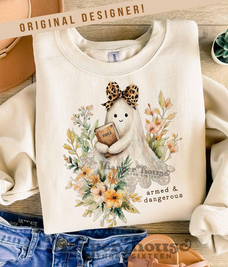 May include: Cream-colored sweatshirt with a graphic of a ghost holding a Bible, wearing a leopard print bow, and surrounded by floral elements. Text on the sweatshirt reads "armed & dangerous" and "Thanker House Sixteen". The text "ORIGINAL DESIGNER!" is also visible.