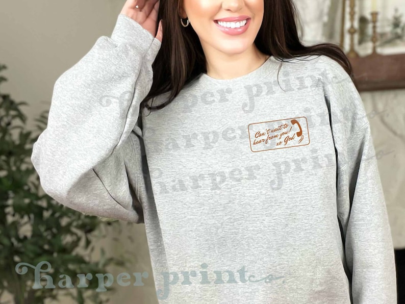 May include: A gray sweatshirt with a brown pocket featuring a phone graphic and the text "Can't wait to hear from you! So God"