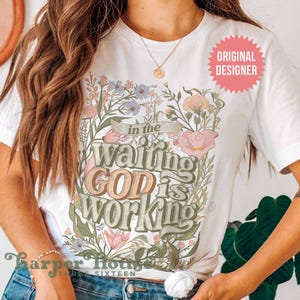 May include: White t-shirt with a floral design and the text "in the waiting GOD is WORKING".