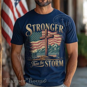 May include: A navy blue t-shirt with a distressed American flag and a wooden cross. The text "Stronger Than the Storm" is printed on the shirt.