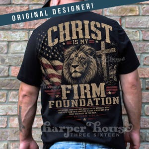 May include: Black t-shirt featuring a lion's head, American flag, and a cross. The text reads "CHRIST IS MY FIRM FOUNDATION" with a Bible verse. The shirt has a distressed, vintage look.