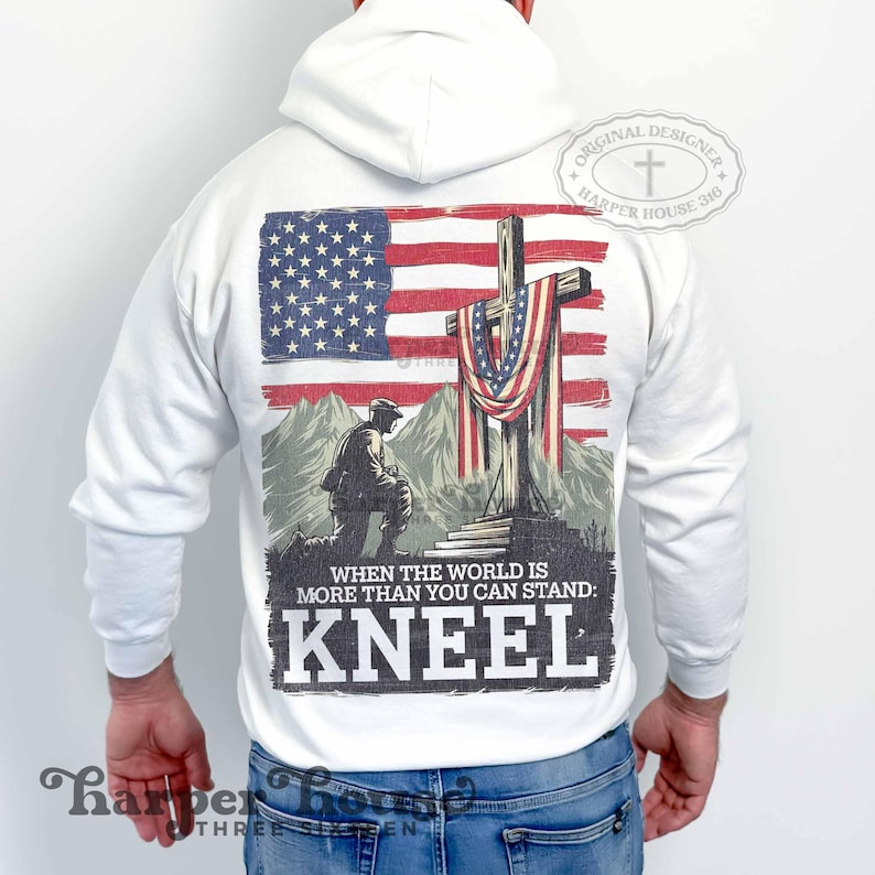 Pode incluir: Camisola de capuz branca com uma estampa gr&aacute;fica de um soldado ajoelhado em frente a uma cruz com uma bandeira americana drapeada sobre ela. O texto "WHEN THE WORLD IS MORE THAN YOU CAN STAND: KNEEL" est&aacute; impresso abaixo do gr&aacute;fico. O texto "ORIGINAL DESIGNER HARPER HOUSE 316" est&aacute; impresso em um c&iacute;rculo na parte superior da camisola.