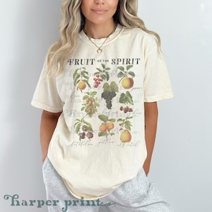 Peut inclure: Un t-shirt à manches courtes de couleur crème avec le texte "FRUIT OF THE SPIRIT" en haut. Le motif présente diverses illustrations de fruits, notamment des raisins, des pêches, des poires et des baies, avec des mots en cursive sous chaque fruit.