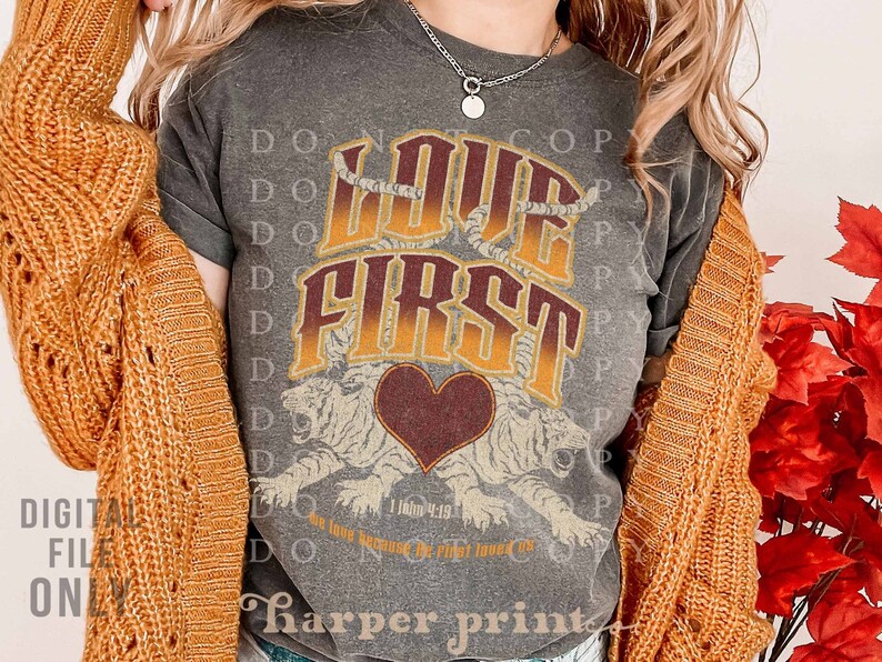 May include: A gray t-shirt with a vintage-style graphic that says "Love First" with a heart and tiger design. The text "I John 4:19" is also included on the shirt.