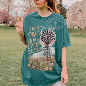 May include: A teal blue t-shirt featuring a vintage-style graphic of a windmill and flowers. The text on the shirt reads "I will praise him in every season" and "Psalm 34:1".
