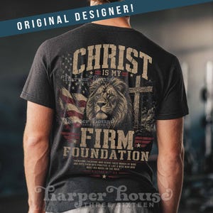 May include: Dark gray t-shirt featuring a lion's head, cross, and American flag graphic. Text includes "Christ is my Firm Foundation" and a Bible verse. The shirt is from Harper House.