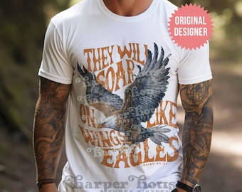May include: A white short-sleeve t-shirt featuring a graphic of a flying eagle in the center. The text reads "THEY WILL SOAR ON WINGS LIKE EAGLES" in orange, with "ISAIAH 40:31" below. A pink starburst badge in the corner says "ORIGINAL DESIGNER".