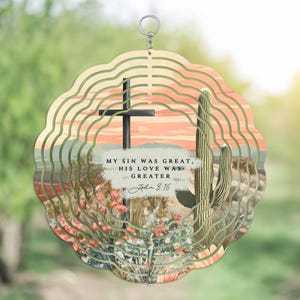 May include: A metal wind chime with a desert sunset design. The chime features a cross, cacti, and the text "My sin was great, his love was greater, John 3:16".