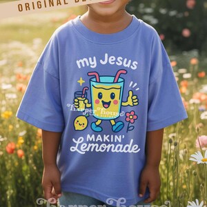 Op de afbeelding: Een lichtblauw t-shirt met de tekst "my Jesus makin' lemonade" en een cartoon limonade karakter. Het shirt heeft ook de tekst "ORIGINAL DESIGNER!" en de merknaam "Harper House Three Sixteen".