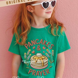 May include: A green t-shirt with the embroidered words "PANCAKES & PRAYER" in a playful font. The design includes a stack of pancakes with syrup, hearts, and floral accents. The shirt is worn by a person wearing heart-shaped sunglasses.
