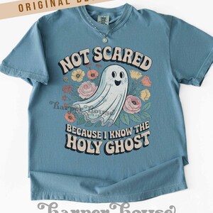 May include: A blue t-shirt with the text "NOT SCARED BECAUSE I KNOW THE HOLY GHOST" and a cartoon ghost surrounded by flowers. The shirt has short sleeves and a round neckline. The words "ORIGINAL DESIGNER!" are at the top.