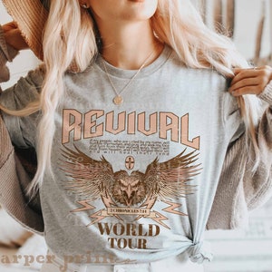 May include: A gray t-shirt with a graphic design featuring an eagle with wings spread, a lightning bolt, and the text "REVIVAL" in pink, "2 CHRONICLES 7:14" in brown, and "WORLD TOUR" in brown.