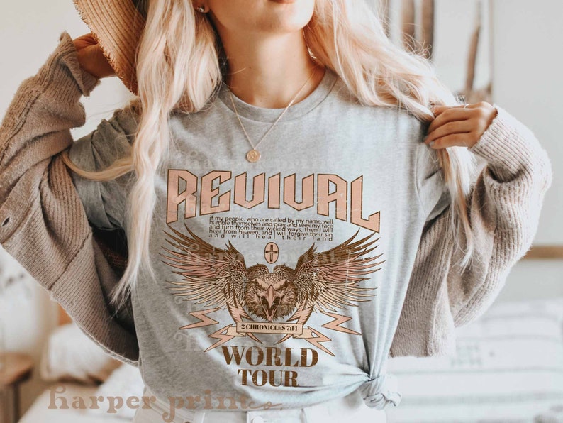 May include: A gray t-shirt with a graphic design featuring an eagle with wings spread, a lightning bolt, and the text "REVIVAL" in pink, "2 CHRONICLES 7:14" in brown, and "WORLD TOUR" in brown.