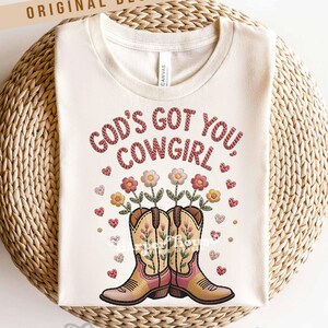 Op de afbeelding: Cr&egrave;mekleurig T-shirt met de tekst "GOD'S GOT YOU COWGIRL" in een gestikte stijl. Het ontwerp omvat geborduurde cowboylaarzen met bloemen en harten. De woorden "ORIGINAL DESIGNER!" staan bovenaan.