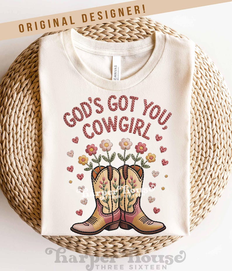 Op de afbeelding: Cr&egrave;mekleurig T-shirt met de tekst "GOD'S GOT YOU COWGIRL" in een gestikte stijl. Het ontwerp omvat geborduurde cowboylaarzen met bloemen en harten. De woorden "ORIGINAL DESIGNER!" staan bovenaan.