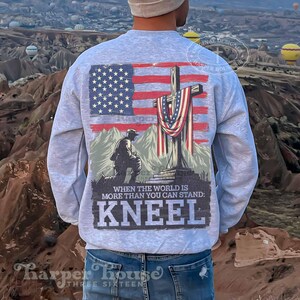 Pode incluir: Uma camisa de moletom cinza com um gr&aacute;fico de um soldado ajoelhado em frente a uma cruz com uma bandeira americana drapeada sobre ela. O texto "When the world is more than you can stand: Kneel" est&aacute; impresso abaixo do gr&aacute;fico.