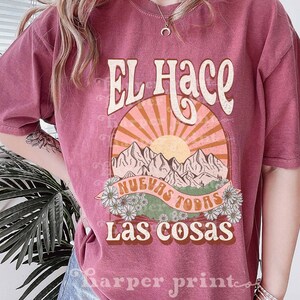 May include: A pink t-shirt with a vintage-style graphic design featuring a sun, mountains, and the text "El Hace Nuevas Todas Las Cosas".