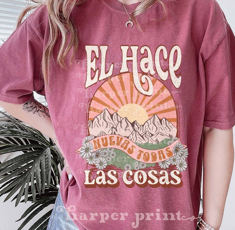 May include: A pink t-shirt with a vintage-style graphic design featuring a sun, mountains, and the text "El Hace Nuevas Todas Las Cosas".