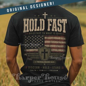 May include: Black t-shirt with a distressed American flag design and the words "HOLD FAST." The shirt includes a cross, stars, and the text "HOLD FAST TO WHAT IS GOOD." Additional text includes "1 THESSALONIANS 5:21" and "DISCERN * HOLD * STAND."