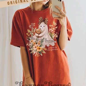 May include: Burnt orange t-shirt featuring a cute ghost design holding a book, surrounded by floral elements. The ghost has a leopard print bow. Text on the shirt includes "Harper House Sixteen" and "armed & dangerous". The shirt also has the text "ORIGINAL DESIGNER!"