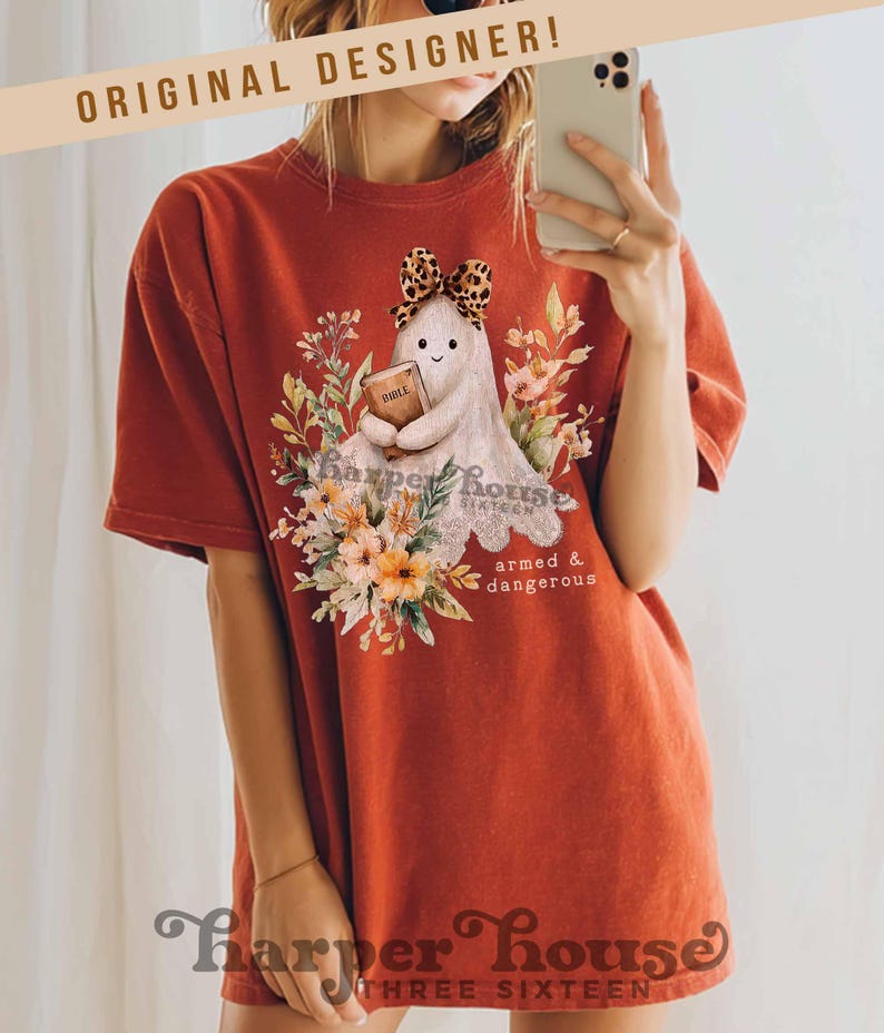 May include: Burnt orange t-shirt featuring a cute ghost design holding a book, surrounded by floral elements. The ghost has a leopard print bow. Text on the shirt includes "Harper House Sixteen" and "armed & dangerous". The shirt also has the text "ORIGINAL DESIGNER!"