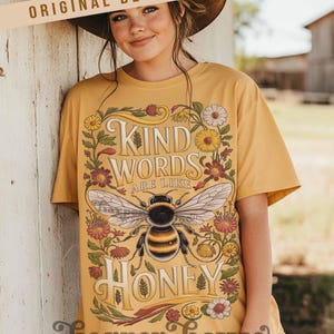 Peut inclure: T-shirt jaune moutarde avec un motif floral et une abeille. Le motif comprend les mots "Kind Words Are Like Honey". Le t-shirt est de Harper House Three Sixteen.