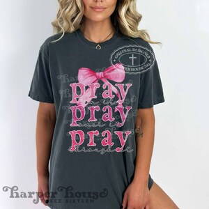 May include: A dark grey t-shirt with a pink ribbon and the text "Pray on it, Pray over it, Pray through it".