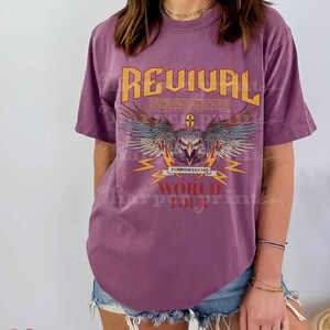 May include: Purple t-shirt with a graphic design of an eagle with wings spread, lightning bolts, and the text "REVIVAL 2 CHRONICLES 7:14 WORLD TOUR".
