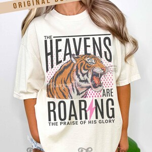 May include: Cream-colored t-shirt featuring a roaring tiger illustration with the text "THE HEAVENS ARE ROARING" and "THE PRAISE OF HIS GLORY". The design includes a pink lightning bolt and a pink dotted background.