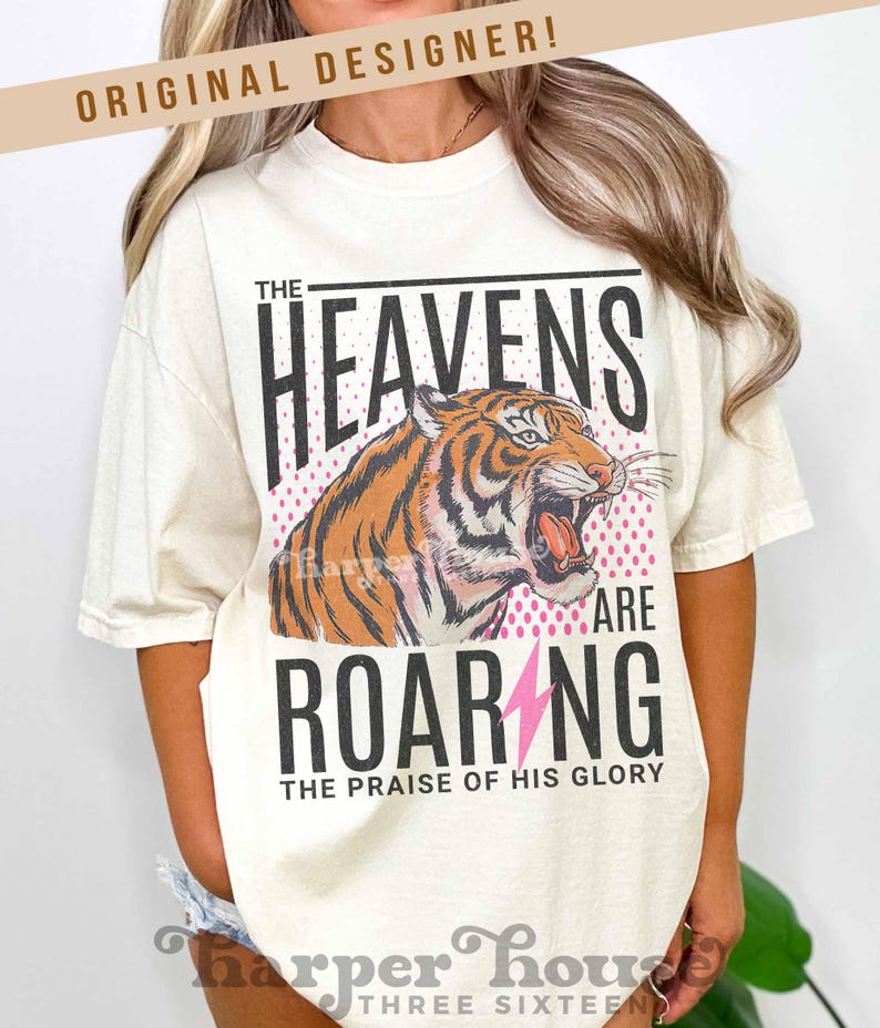 May include: Cream-colored t-shirt featuring a roaring tiger illustration with the text "THE HEAVENS ARE ROARING" and "THE PRAISE OF HIS GLORY". The design includes a pink lightning bolt and a pink dotted background.