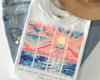 May include: A white t-shirt featuring a graphic of a blue cross standing in a sea under a vibrant sunrise with pink, orange, and blue clouds. The text reads: THE CROSS HAS SPOKEN, I AM FORGIVEN 1 Peter 2:24. The shirt is displayed with blue jeans and a tan hat.
