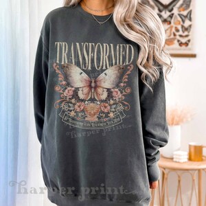 May include: A dark gray sweatshirt with a vintage-style graphic print of a butterfly and floral design. The text "TRANSFORMED" is printed in large letters above the butterfly. The text "BE TRANSFORMED BY THE RENEWING OF YOUR MIND" is printed below the butterfly.