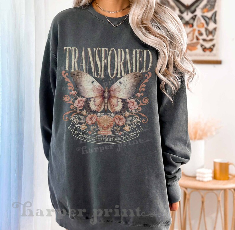 May include: A dark gray sweatshirt with a vintage-style graphic print of a butterfly and floral design. The text "TRANSFORMED" is printed in large letters above the butterfly. The text "BE TRANSFORMED BY THE RENEWING OF YOUR MIND" is printed below the butterfly.