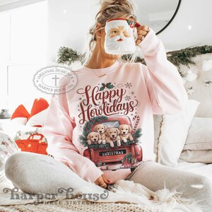 Op de afbeelding: Roze trui met een rode vrachtwagen die een groep golden retriever-puppies vervoert. De vrachtwagen heeft de tekst "Christmas" op de achterkant. De trui heeft de tekst "Happy Howlidays" in rode letters.