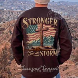 May include: A brown sweatshirt with a graphic print of an American flag with a wooden cross in front of it. The text "Stronger Than the Storm" is printed below the flag. The sweatshirt has a crew neck and long sleeves.
