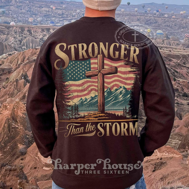 May include: A brown sweatshirt with a graphic print of an American flag with a wooden cross in front of it. The text "Stronger Than the Storm" is printed below the flag. The sweatshirt has a crew neck and long sleeves.