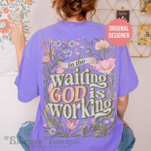 May include: Purple t-shirt with a floral design and the text "in the waiting GOD is working".