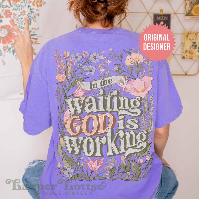 May include: Purple t-shirt with a floral design and the text "in the waiting GOD is working".