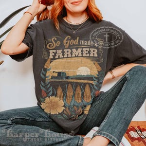 Op de afbeelding: Een antracietgrijs T-shirt met korte mouwen en een grafische print met de tekst So God made a FARMER en Original Designer Harper House 316. Het ontwerp toont een tractor in een veld, maïskolven en een grote zon.