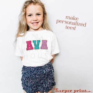 May include: A young girl wearing a white t-shirt with the name "AVA" in colorful letters. The shirt is personalized with a gold glitter outline. The girl is wearing blue and white polka dot shorts.